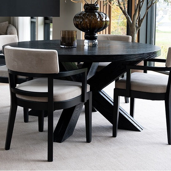 Round black wooden dining table with black wooden & taupe velvet padded dining chairs with armrests.
